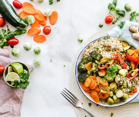 A healthy grain bowl filled with quinoa and other vegetables is on a table next to a fork, surrounded by fresh ingredients like zucchini, tomatoes, ginger, and lime.