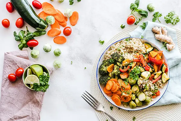 A healthy grain bowl filled with quinoa and other vegetables is on a table next to a fork, surrounded by fresh ingredients like zucchini, tomatoes, ginger, and lime.
