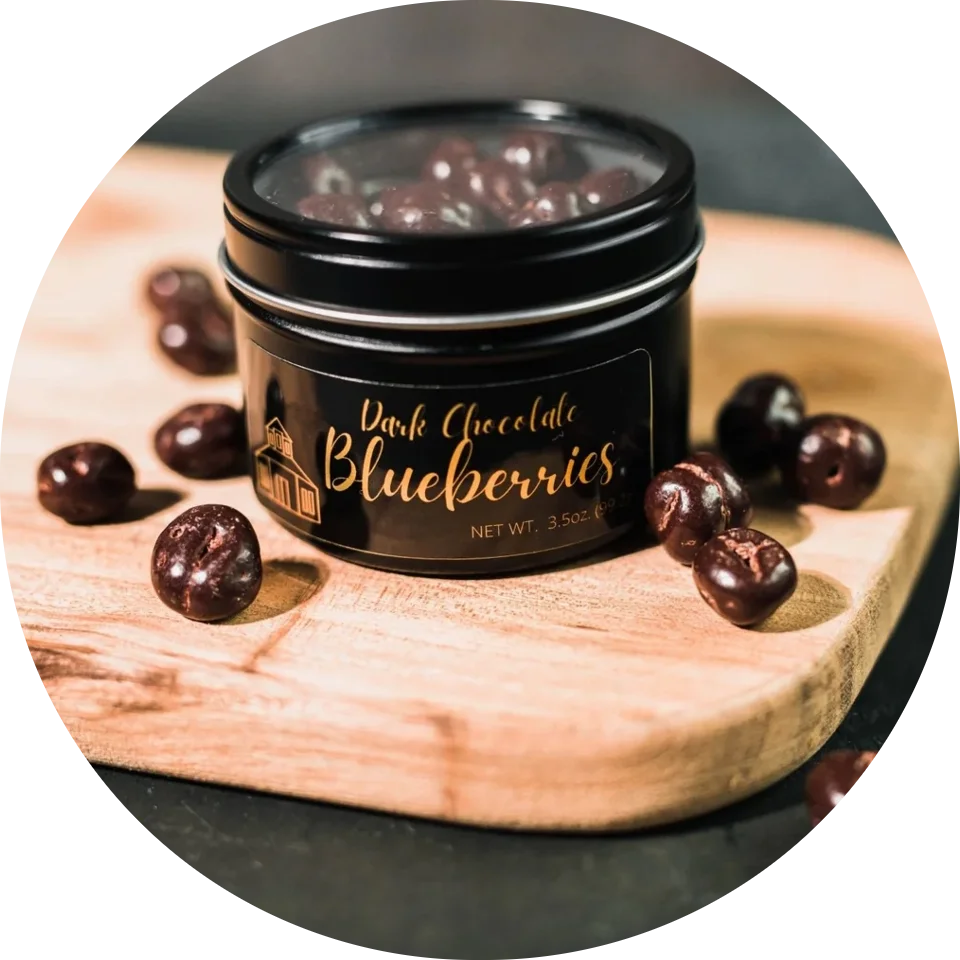 Dark Chocolate Blueberries tin with chocolate-coated blueberries scattered on a wooden board