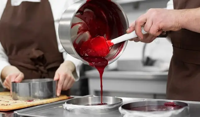 Confectioners pouring bright red berry filling from a saucepan into cake pan in commercial kitchen