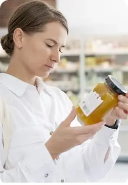 A consumer is examining a jar of food in a grocery store, carefully reading the product label for nutritional or ingredient information.