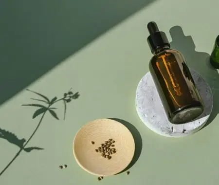 CBD hemp oil dropper bottle and seeds with cannabis plant shadow on sage green surface