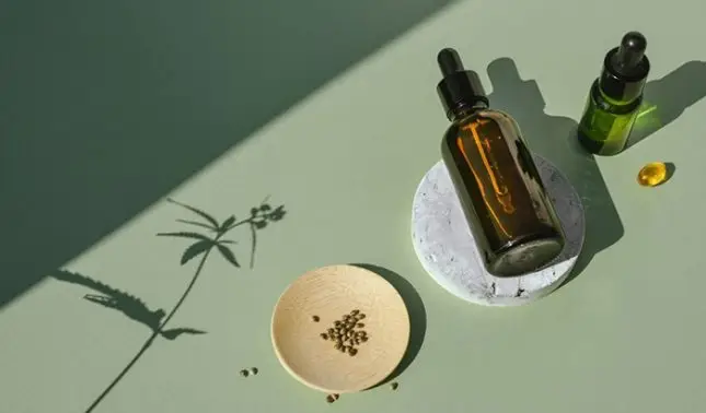 CBD hemp oil dropper bottle and seeds with cannabis plant shadow on sage green surface