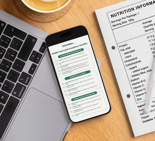 Workspace showing laptop, smartphone displaying FSANZ food label compliance checklist, printed nutrition information panel, and coffee cup on wooden desk